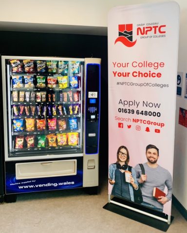 Vending machine with snacks and drinks next to an NPTC promotional stand.